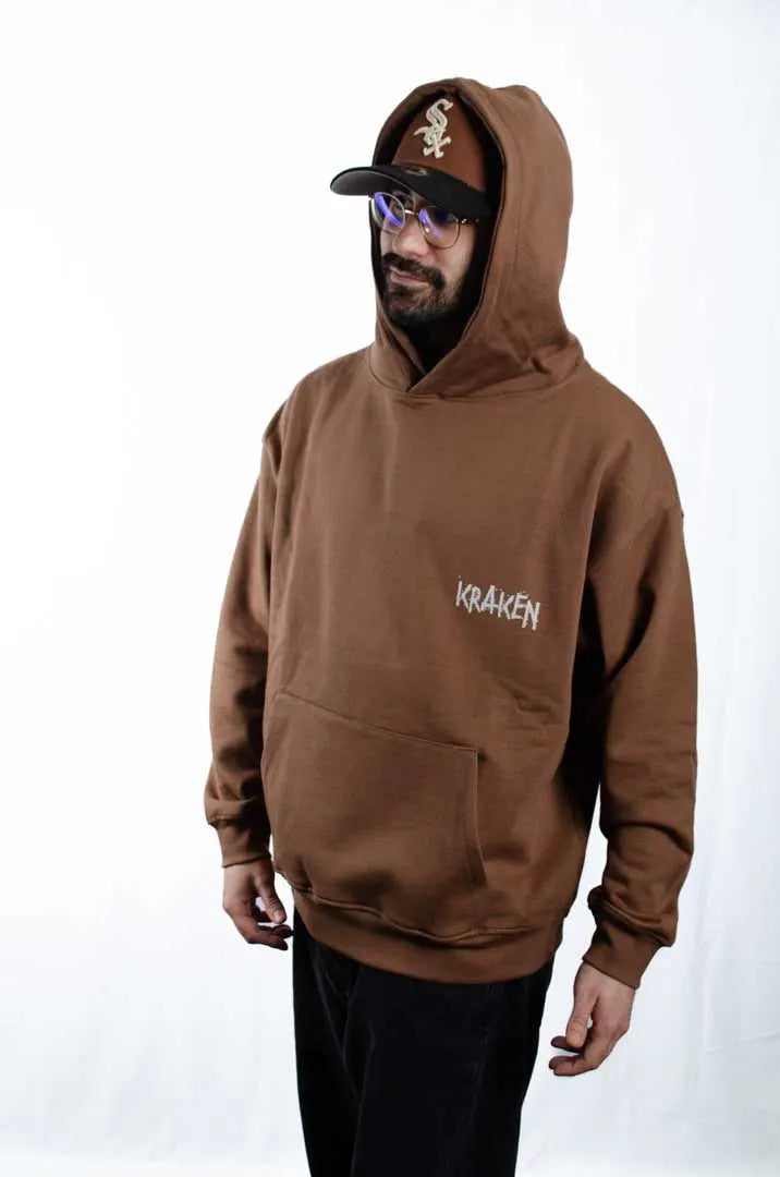 Vista lateral de un modelo hombre vistiendo la Sudadera El Orgullo de tu Papá. Sudadera streetwear oversized con capucha, en color Brownie especial y fabricada con material de felpa perchada 100% algodón.